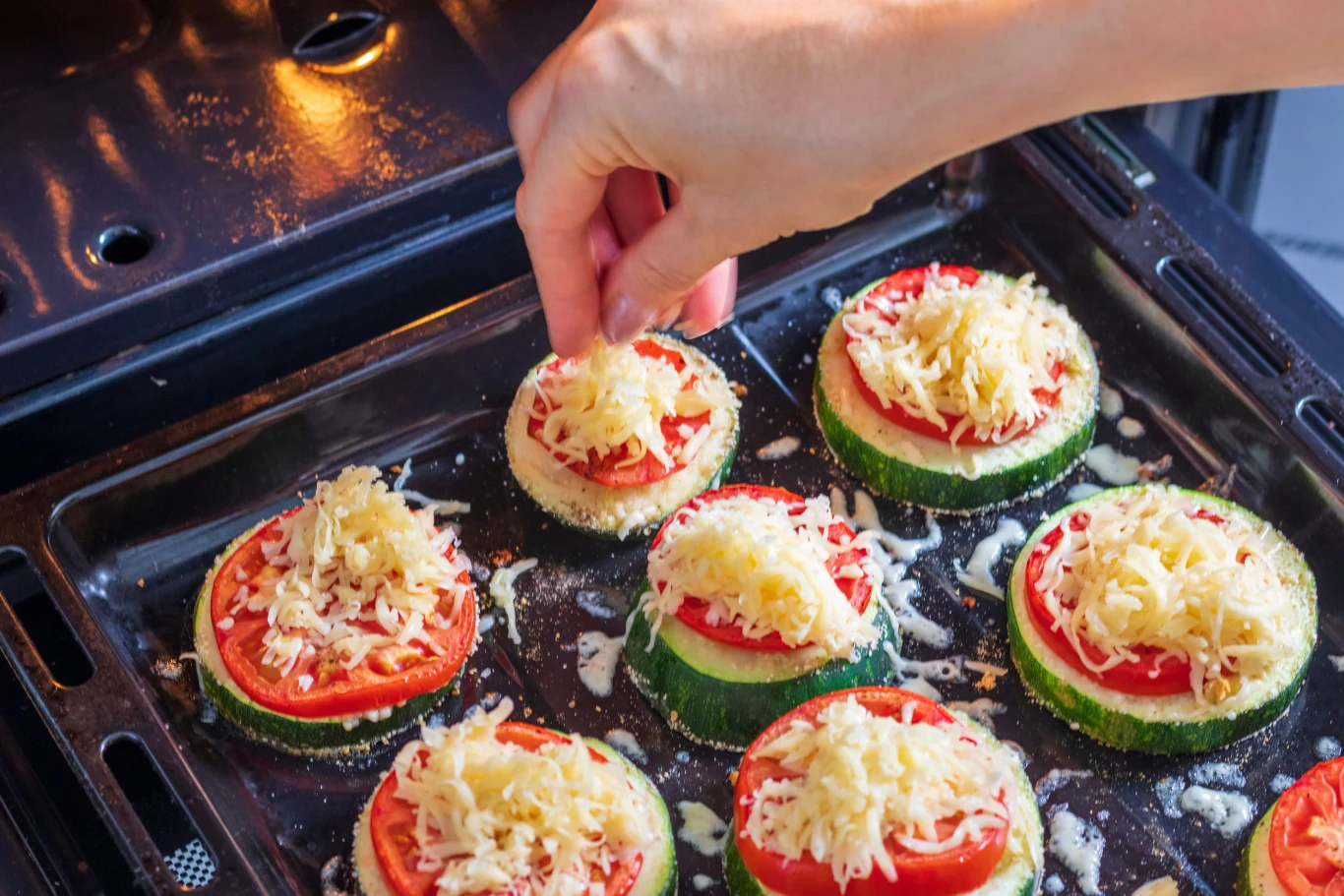 Zapiekam kilka składników na plastrach cukinii i mam zdrową przekąskę Pizzerinki z cukinii z pomidorem i tartym serem ułożone na blasze, przygotowane do zapiekania w piekarniku.