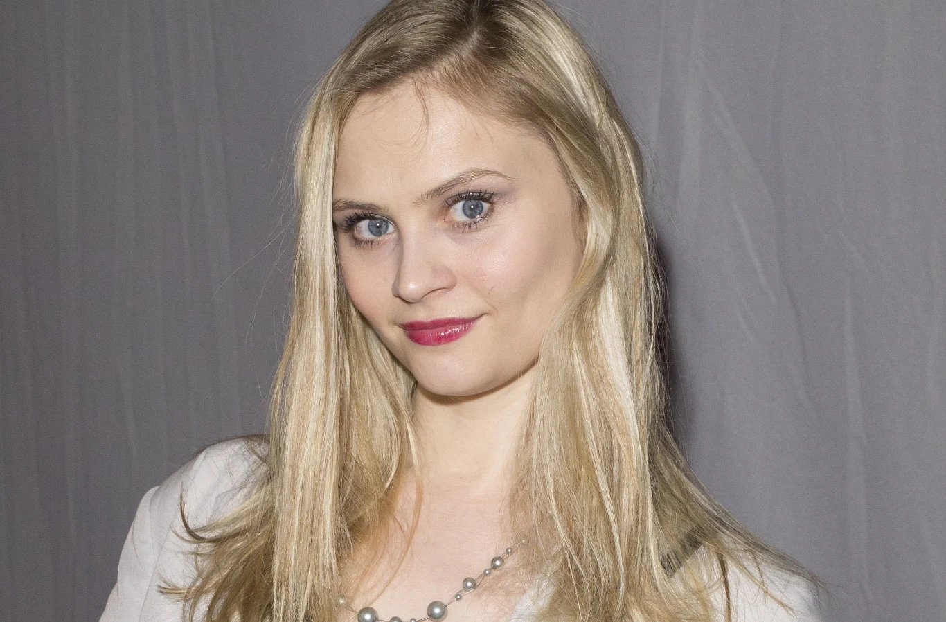 Patrycja Szczepanowska Młoda kobieta o długich blond włosach, ubrana w jasną bluzkę i naszyjnik z pereł, patrząca bezpośrednio w obiektyw na tle szarej zasłony.