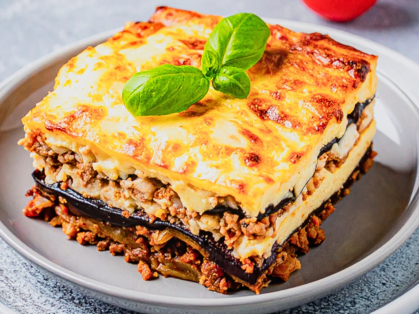 Tradycyjna grecka moussaka to połączenie bakłażana, mięsa wołowego i beszamelu. Kawałek greckiej moussaki z warstwami bakłażana, mięsa wołowego i beszamelu, udekorowany świeżą bazylią.