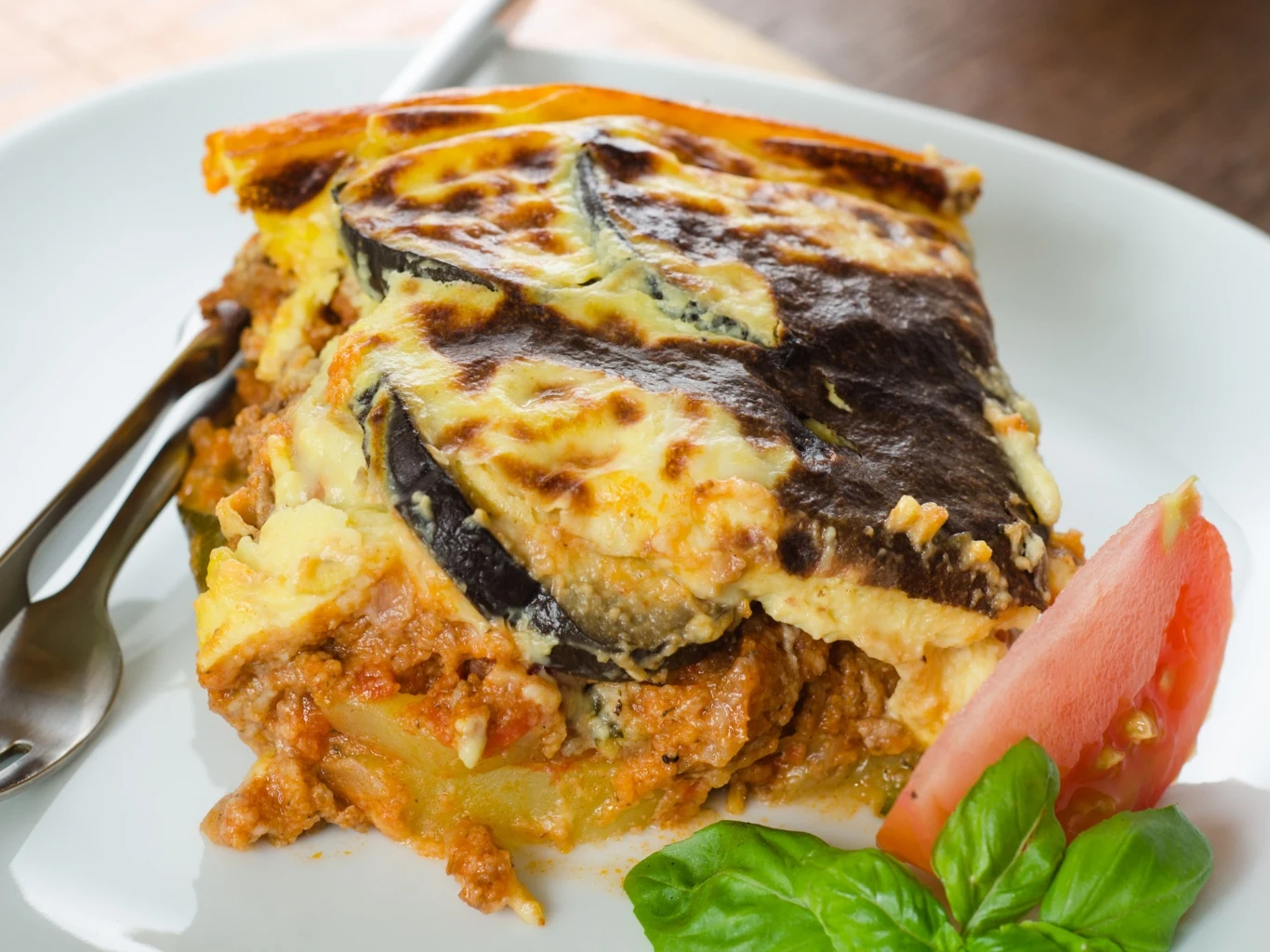 Moussaka jest lepsza od lazani. Porcja zapiekanki z warstwami bakłażana, mięsa mielonego i sosu beszamelowego, podana z kawałkiem pomidora i listkami bazylii.