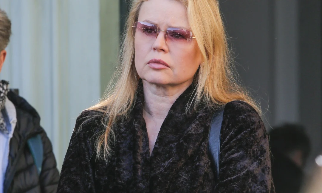Magdalena Wójcik Kobieta z blond włosami w okularach przeciwsłonecznych i ciemnym futrzanym płaszczu stoi w jasnym świetle dziennym, z poważnym wyrazem twarzy.