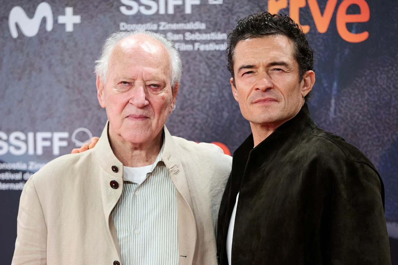 Werner Herzog i Orlando Bloom w San Sebastian Dwóch elegancko ubranych mężczyzn pozuje ramię w ramię na tle ścianki z logo wydarzenia filmowego.