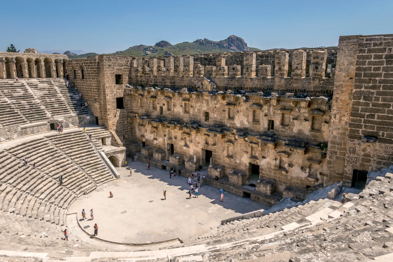 Teatr w Aspendos warto odwiedzić zarówno za dnia, jak i podczas wieczornych wydarzeń Starożytny kamienny amfiteatr z półkolistymi rzędami siedzeń, dużą monumentalną sceną i masywnymi murami otaczającymi widownię, z pojedynczymi osobami spacerującymi po arenie.