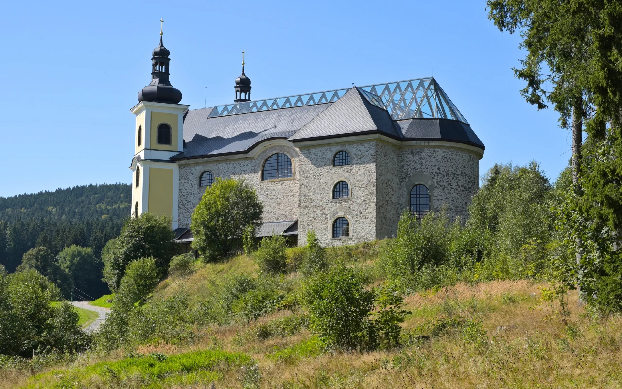 Kamienny kościół z dwiema wieżami i charakterystycznym metalowym szkieletem dachu, położony na wzgórzu otoczonym zielenią i lasami.