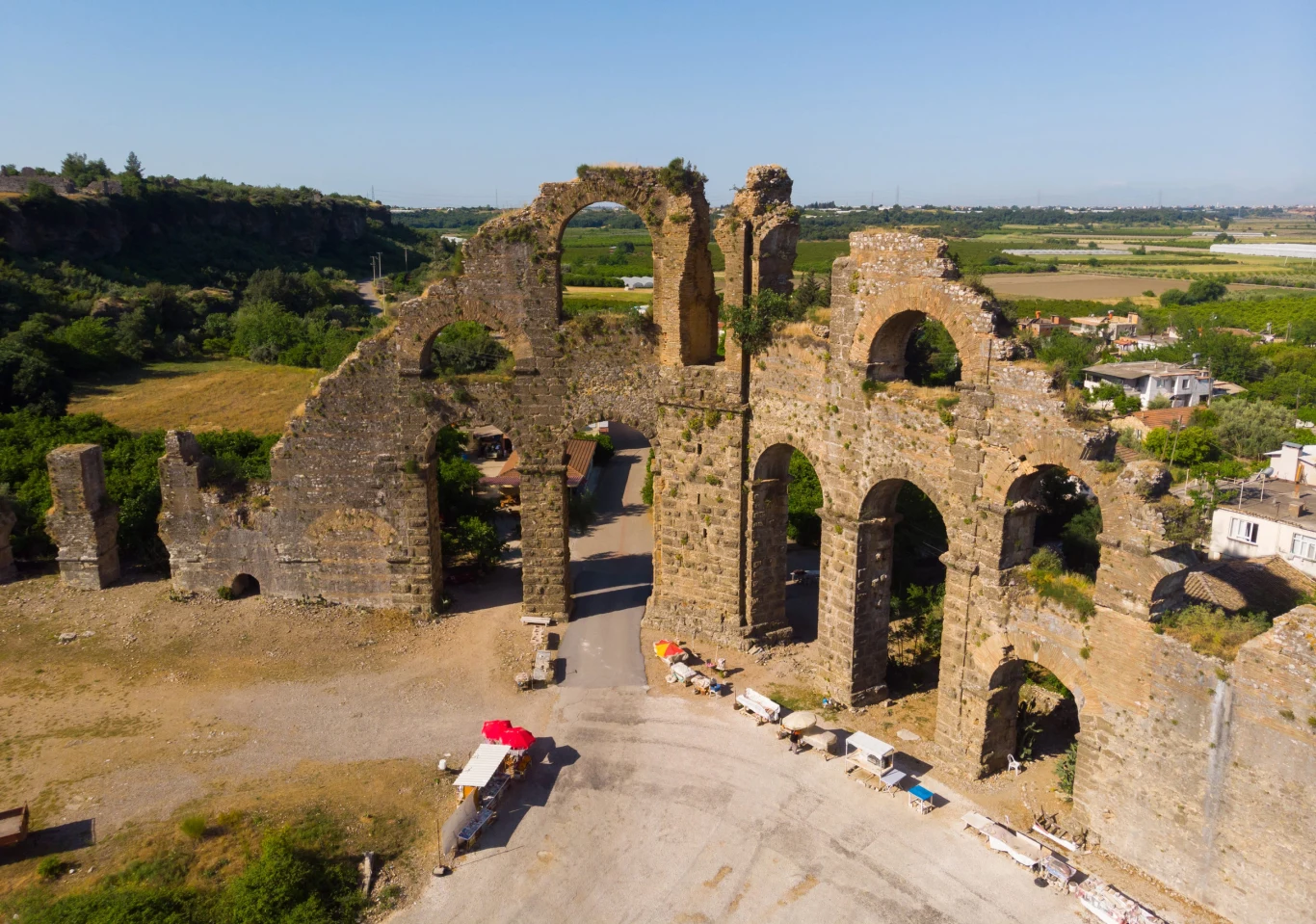 W Turcji przenieś się znad basenu do starożytnego świata Monumentalne ruiny akweduktu z łukowatymi przęsłami, otoczone zielenią i polami uprawnymi, u stóp których rozstawione są kramy z daszkami w kolorze czerwonym i białym.