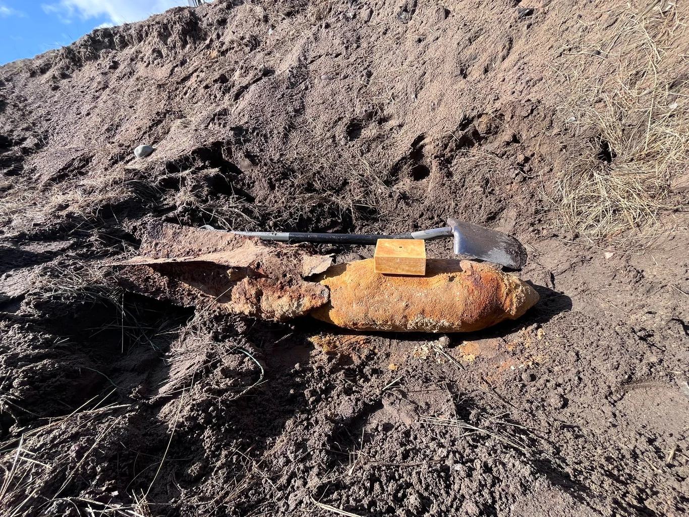 Odnaleziony pocisk tuż przed detonacją Duża, zardzewiała bomba częściowo zakopana w ziemi, obok której leży drewniany klocek i metalowy łom.