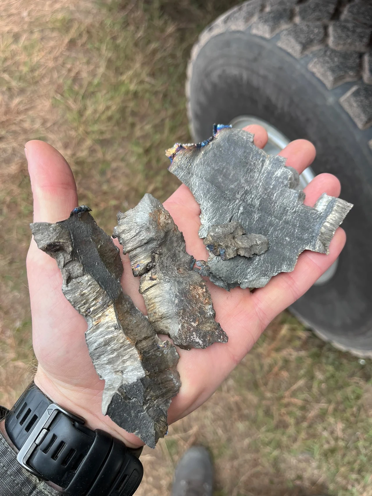 Odłamki pocisku odnalezione po wybuchu Trzy duże, nieregularne kawałki połyskującego metalu spoczywające na otwartej dłoni, w tle fragment terenowej opony i trawa.