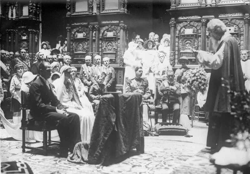 Para młoda siedząca przed ołtarzem podczas ceremonii ślubnej w otoczeniu dostojników i gości ubranych w eleganckie stroje, za którymi znajdują się bogato zdobione, drewniane elementy wystroju kościoła.