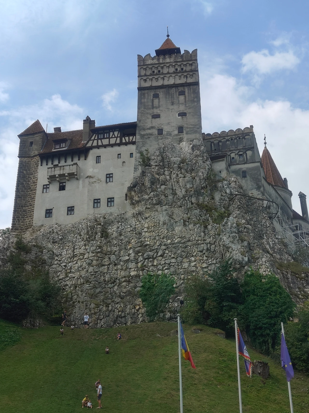Kamienny zamek z wysoką wieżą na skalistym wzgórzu, otoczony zielenią i flagami na pierwszym planie.