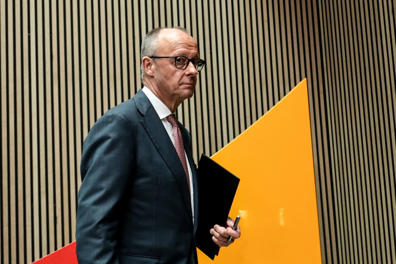 Kanclerz Niemiec Friedrich Merz Elegancko ubrany mężczyzna w garniturze i okularach trzyma czarną teczkę oraz długopis, idąc na tle geometrycznych kształtów w nowoczesnym wnętrzu.