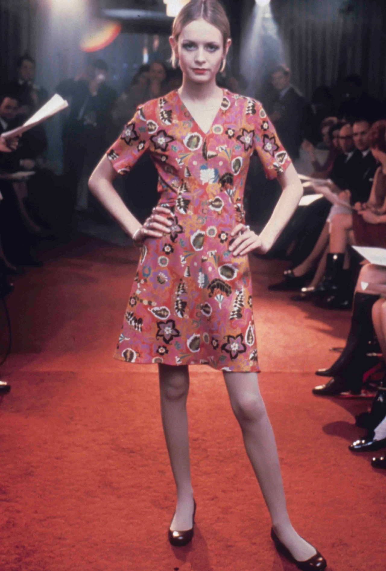 Twiggy Lawson Modelka w krótkiej sukience mini z kolorowym, kwiatowym wzorem na czerwonym dywanie, stojąca w pewnej pozie z rękami na biodrach, wokół publiczność siedząca po obu stronach wybiegu.