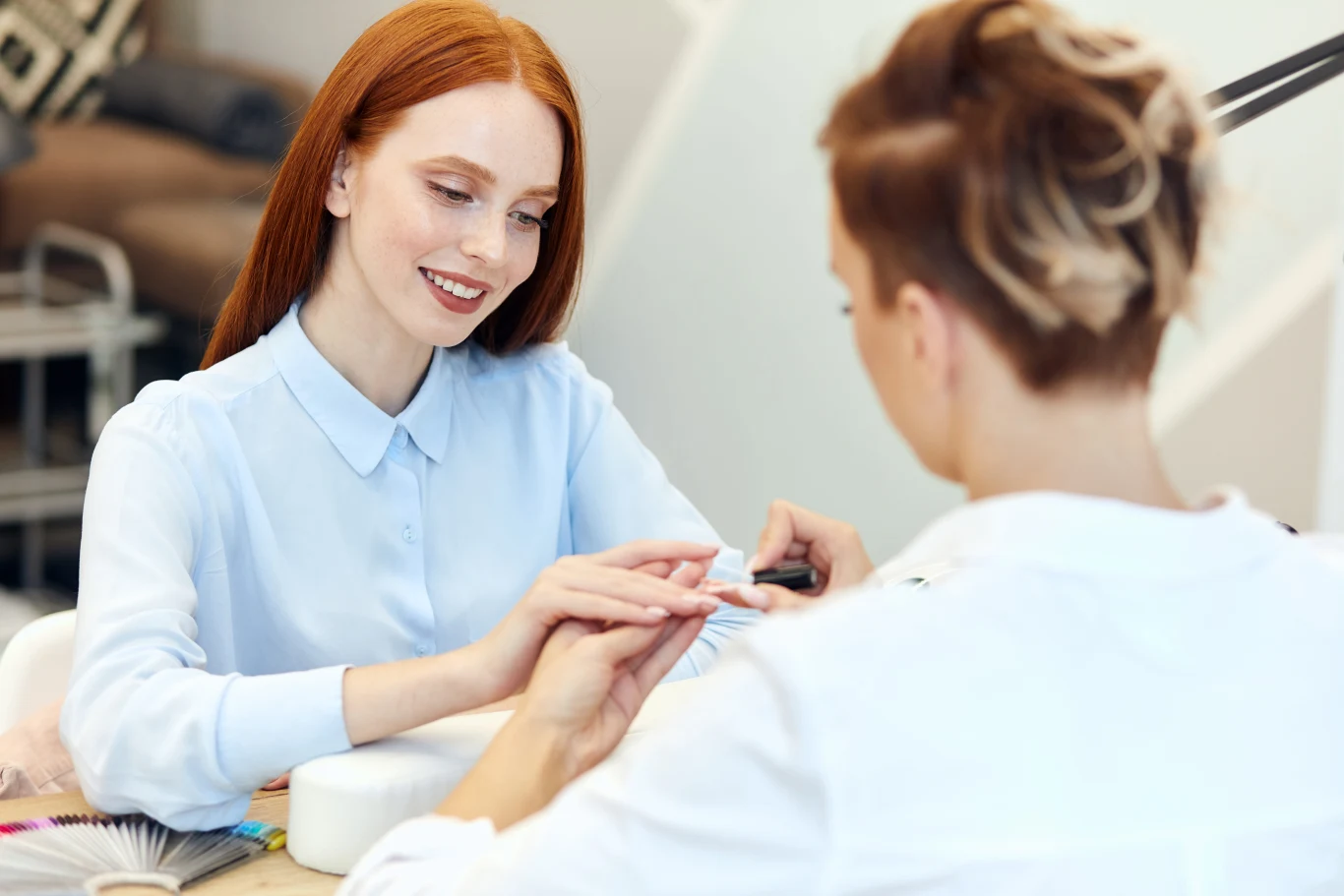 Najmodniejszy manicure na jesień 2026 to pattern french manicure. Sprawdź, czym się wyróżnia. Kobieta o rudych włosach uśmiecha się podczas zabiegu manicure. Najmodniejszy manicure na jesień.