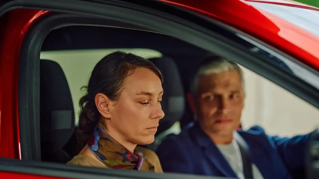 Kadr z serialu "Na Wspólnej" Kadr z serialu Na Wspólnej: kobieta na miejscu pasażera w czerwonym aucie, obok niej kierowca, mężczyzna.
