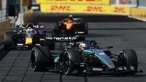 Boliden wyścigowe Formuły 1 rywalizujące ze sobą na torze, na prowadzeniu samochód zespołu Mercedes z widocznym logo Petronas, za nim pojazdy Red Bull Racing i McLarena, w tle wyraźne banery reklamowe DHL.