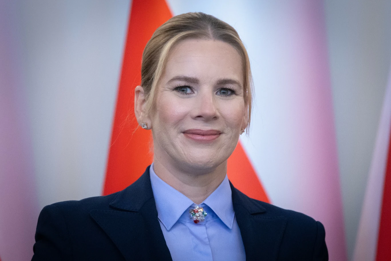 Marta Nawrocka kocha sport Marta Nawrocka w ciemnej marynarce i jasnoniebieskiej koszuli z broszką, na tle czerwono-białych flag.