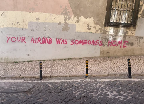 Różowe graffiti z napisem „YOUR AIRBNB WAS SOMEONES HOME” namalowane na jasnej, zniszczonej ścianie budynku, powyżej brukowanego chodnika i ulicy z trzema czarnymi słupkami ostrzegawczymi.