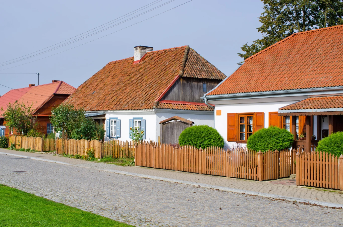 Rząd tradycyjnych domów o bielonych ścianach i czerwonych dachach z widocznymi drewnianymi ogrodzeniami oraz zadbanymi krzewami przed wejściami, położonych przy brukowanej ulicy.