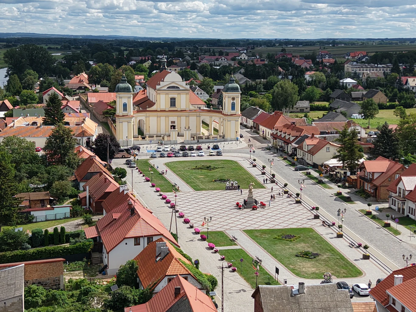 Rynek z zielonym skwerem otoczonym brukowanymi uliczkami, w centralnej części okazały barokowy kościół z dwiema wieżami, po bokach niskie, czerwono-dachowe kamienice, spacerujący ludzie oraz zaparkowane auta przed świątynią.