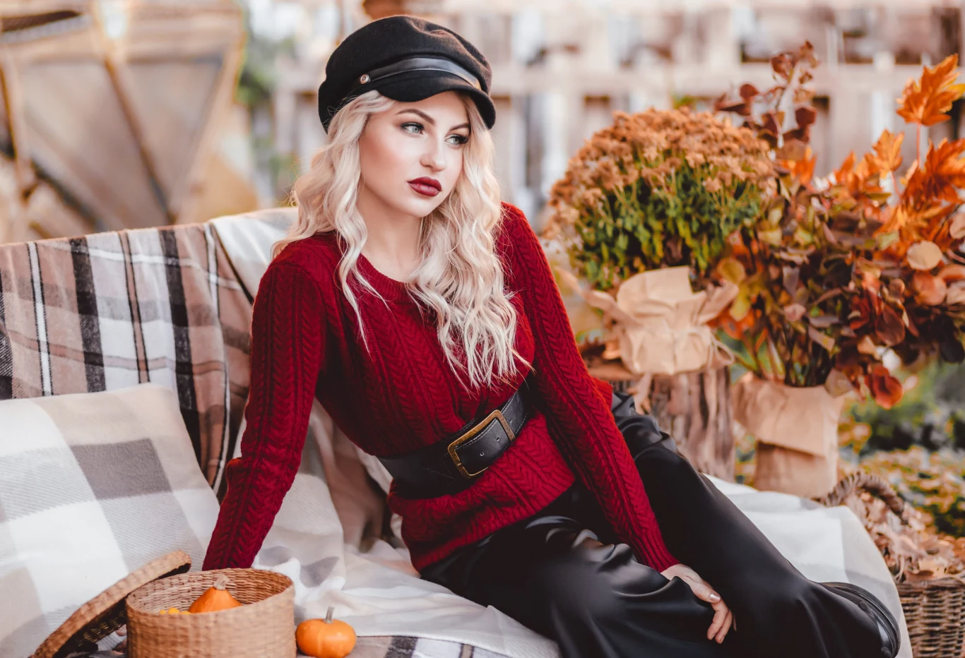 Jesienna równonoc nie będzie dla nich obojętna Kobieta z długimi blond włosami ubrana w czerwony sweter i czarną czapkę z daszkiem, siedząca na kanapie przykrytej kocem, obok niej koszyk z miniaturowymi dyniami, w tle jesienne liście i bukiety suszonych kwiatów.