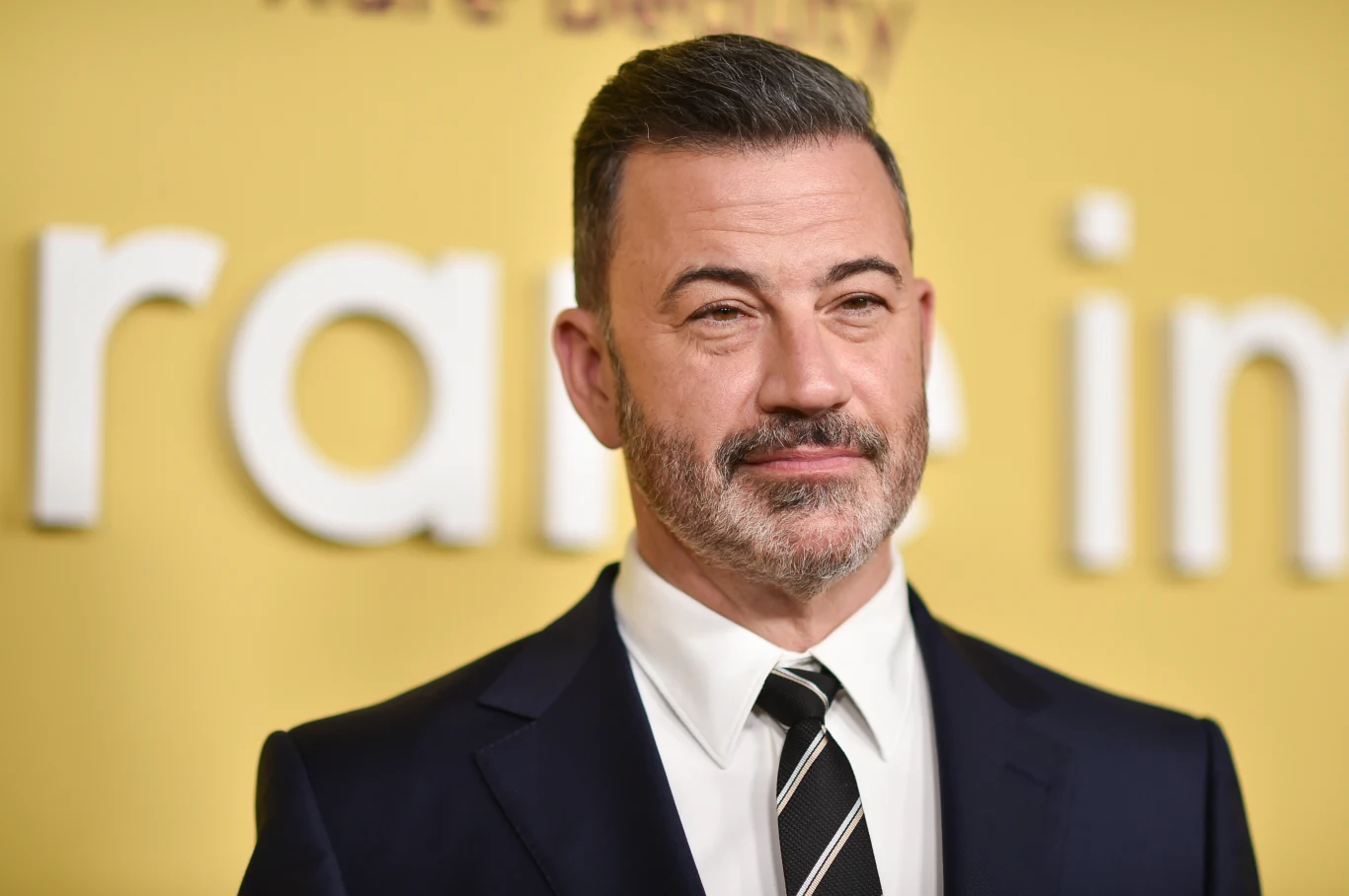 Jimmy Kimmel w eleganckim garniturze z białą koszulą i krawatem w czarne paski, z zarostem i krótkimi, siwymi włosami, stoi na tle żółtej ściany z dużymi, białymi literami.