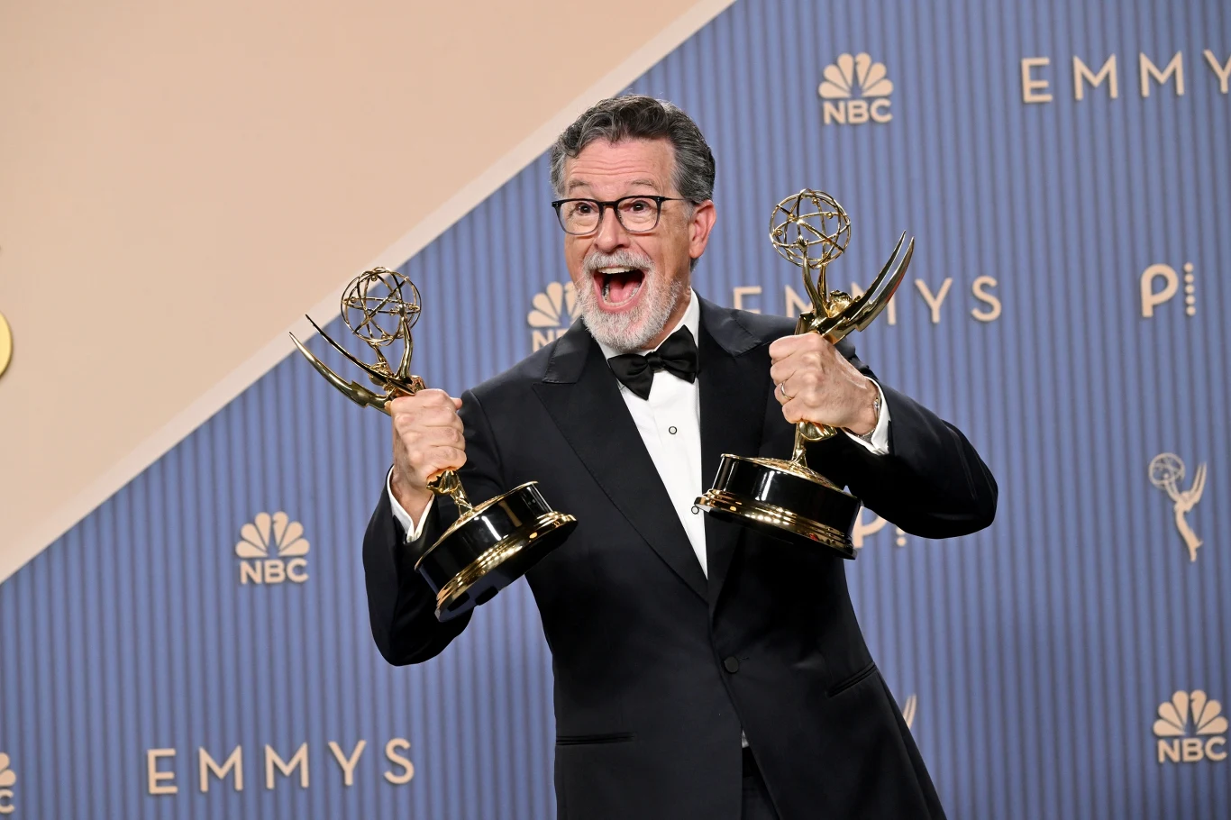 Program Stephena Colberta zniknął z CBS, ale otrzymał za niego nagrody Emmy Mężczyzna w garniturze i muszce trzyma w obu rękach dwie statuetki Emmy, uśmiecha się szeroko stojąc na tle z logo gali.