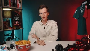 Mężczyzna w jasnej koszuli siedzący przy stole z miską nachosów Doritos, szklanką wody i miniaturowym modelem bolidu, w tle regał z książkami oraz zielona koszulka zespołu wyścigowego podświetlona na czerwono.