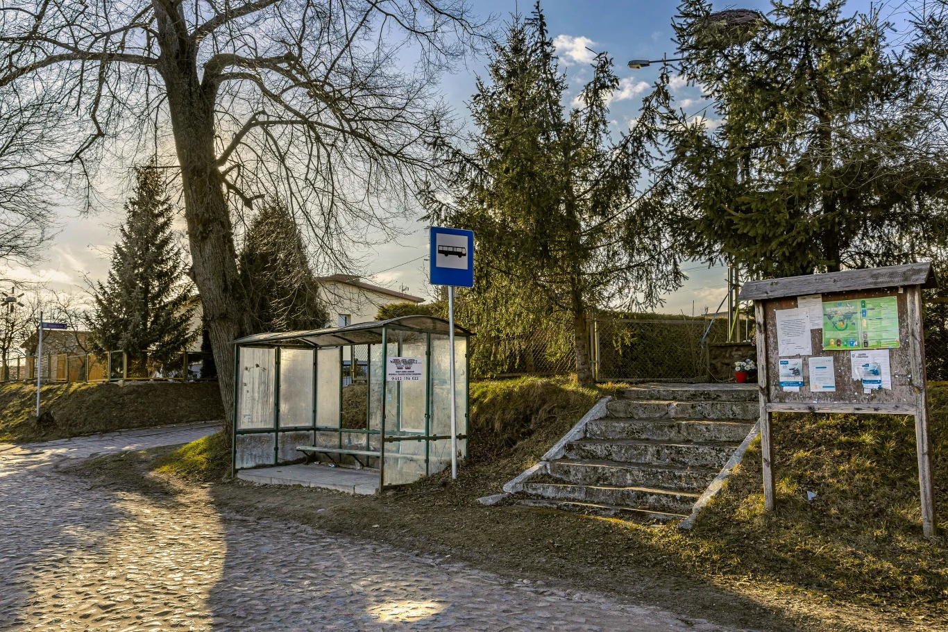 Nawet jeśli odnajdziemy przystanek - czy zorientujemy się, dokąd i o której jedzie autobus? Przystanek autobusowy z przezroczystą wiatą, niebieskim znakiem oraz tablicą informacyjną stojącą obok schodów prowadzących na niewielkie wzniesienie otoczone drzewami.
