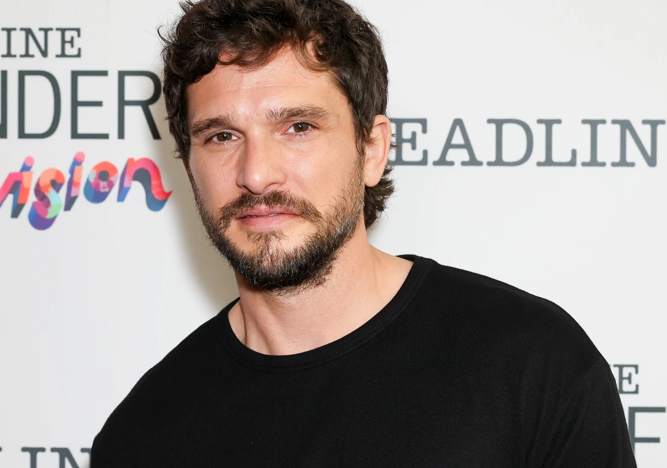 Kit Harington Kit Harington z krótkim, ciemnym zarostem i włosami w czarnym T-shircie na tle ściany z napisami