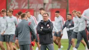 Trener w sportowym stroju z założonymi rękami, otoczony grupą piłkarzy reprezentacji Polski podczas treningu na zielonym boisku.