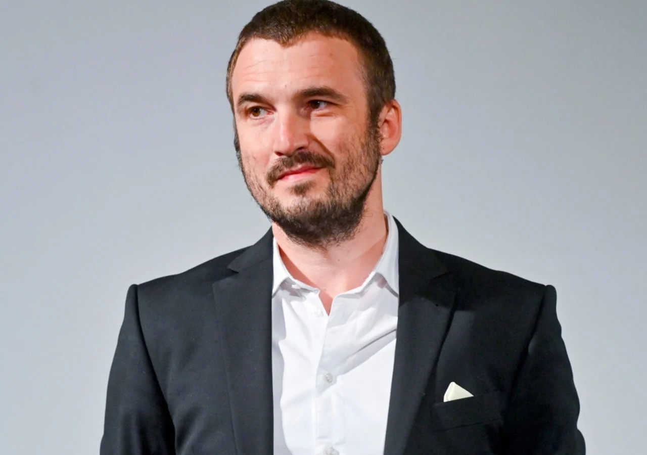 Mężczyzna w ciemnym garniturze z białą koszulą, patrzący lekko w bok, z delikatnym uśmiechem na twarzy.
