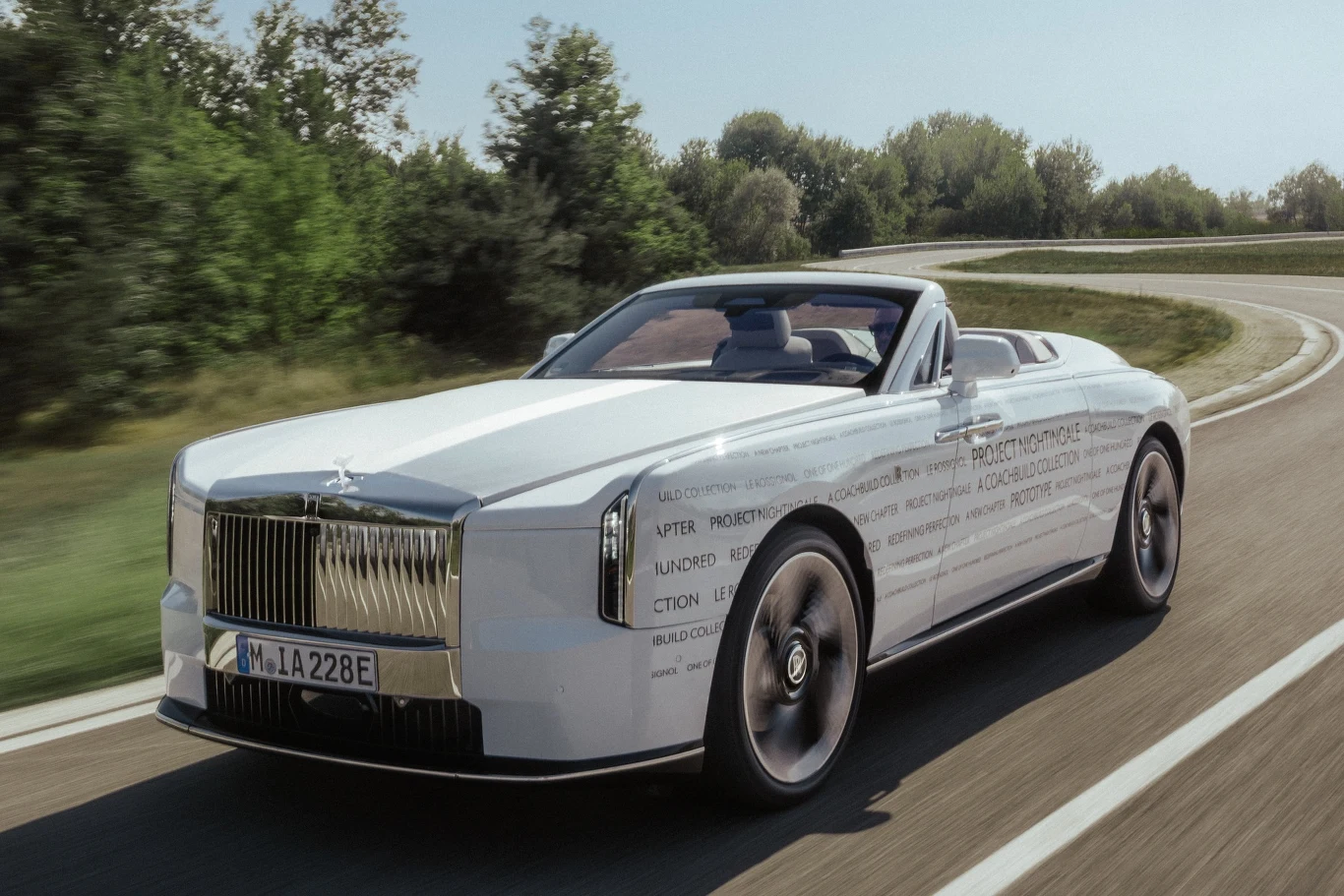 Luksusowy biały kabriolet o nowoczesnym, eleganckim designie porusza się po szerokiej, krętej drodze otoczonej zielenią.