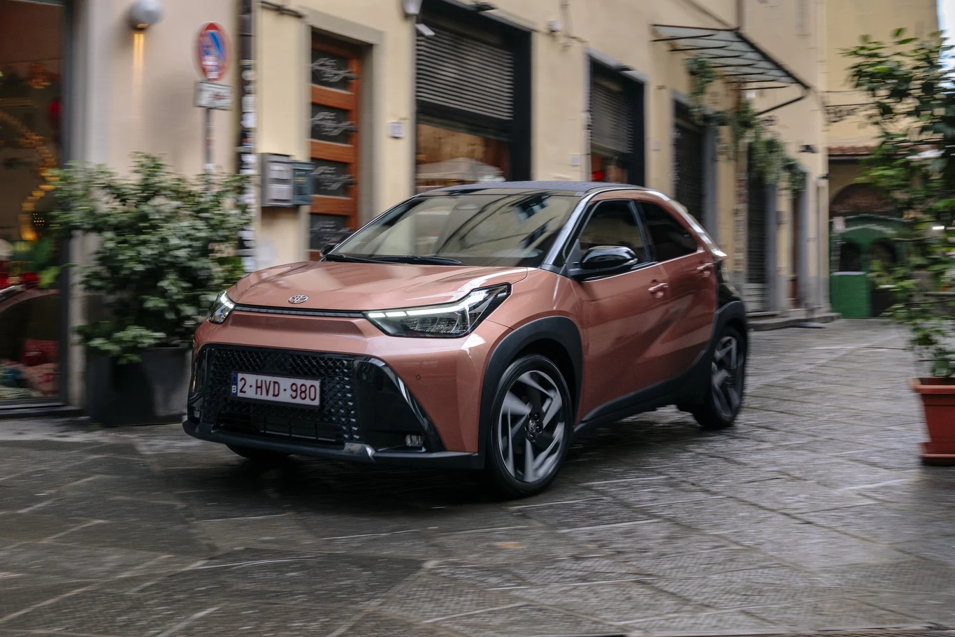 Nowoczesny, miejski crossover Toyota Aygo X w kolorze brązowym jedzie wąską, brukowaną uliczką otoczoną zabudową z witrynami i roślinami doniczkowymi.