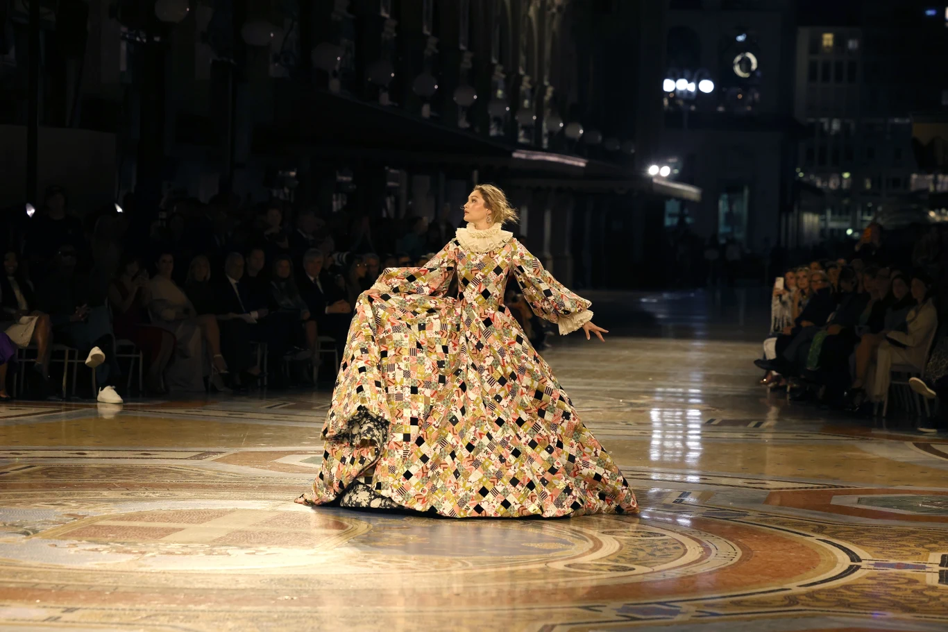 Moda w obecnych czasach nie ma już tylko charakteru użytkowego. To środek przekazu, z którego niezwykle chętnie korzystają artyści Modelka w obszernej, kolorowej sukni z długimi rękawami prezentuje kreację na środku bogato zdobionego holu, otoczona publicznością siedzącą po bokach wybiegu.