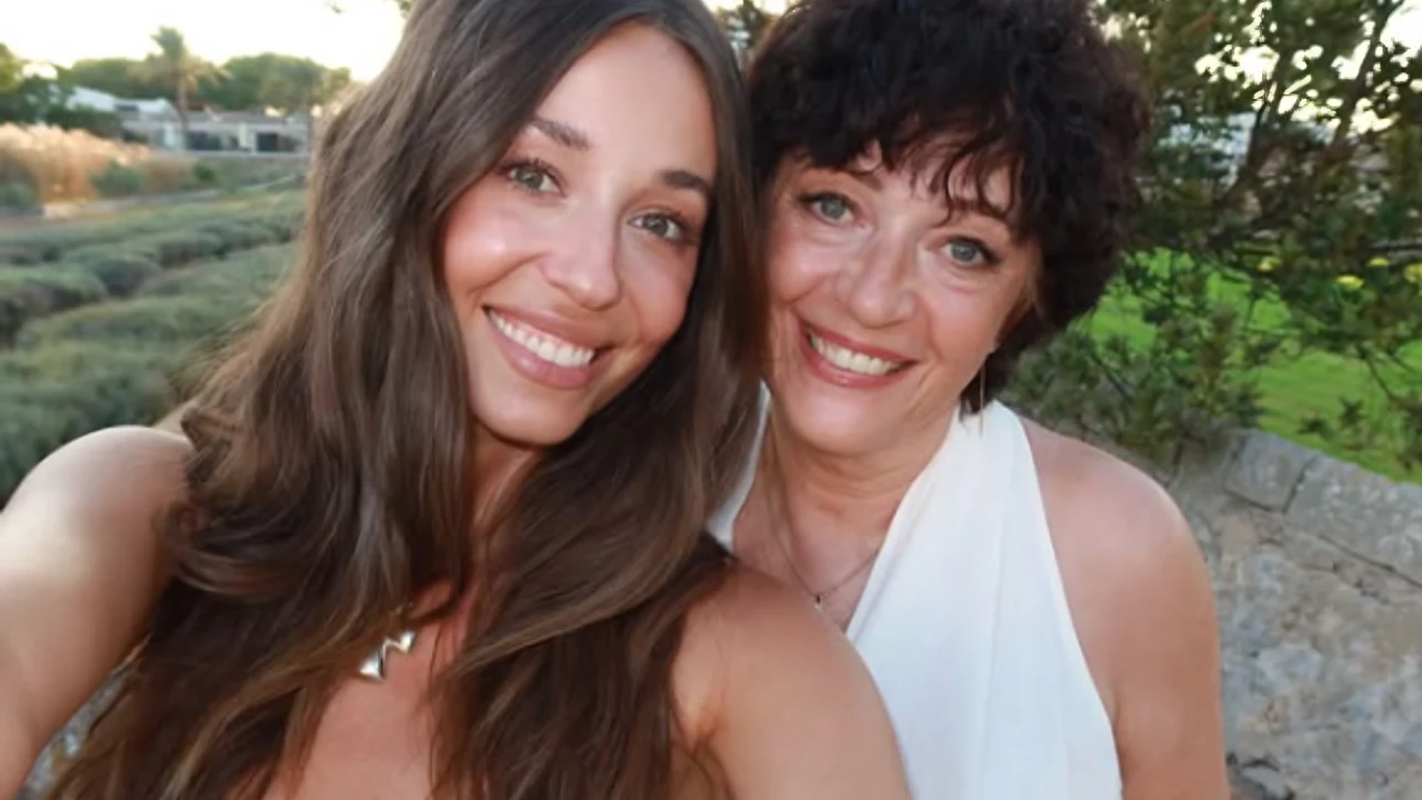 Dwie uśmiechnięte kobiety pozują do selfie na świeżym powietrzu, obie mają na sobie jasne ubrania i wyglądają na zadowolone, w tle widoczna zieleń i kamienny mur.
