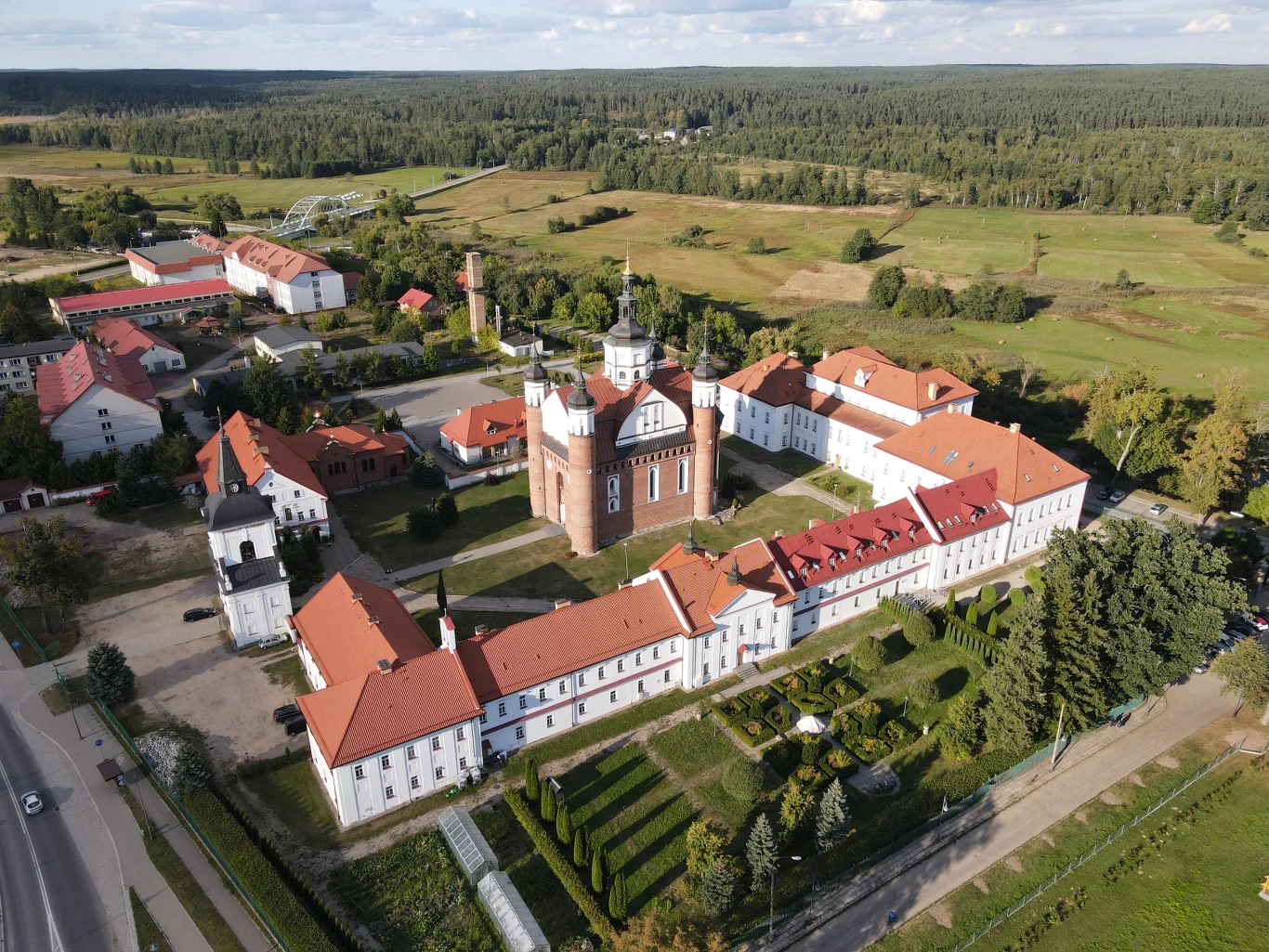 Zabytkowy kompleks klasztorny z charakterystyczną ceglaną wieżą i białymi zabudowaniami z czerwonymi dachami, otoczony uporządkowanym ogrodem i zielenią, w tle rozległe pola oraz lasy.