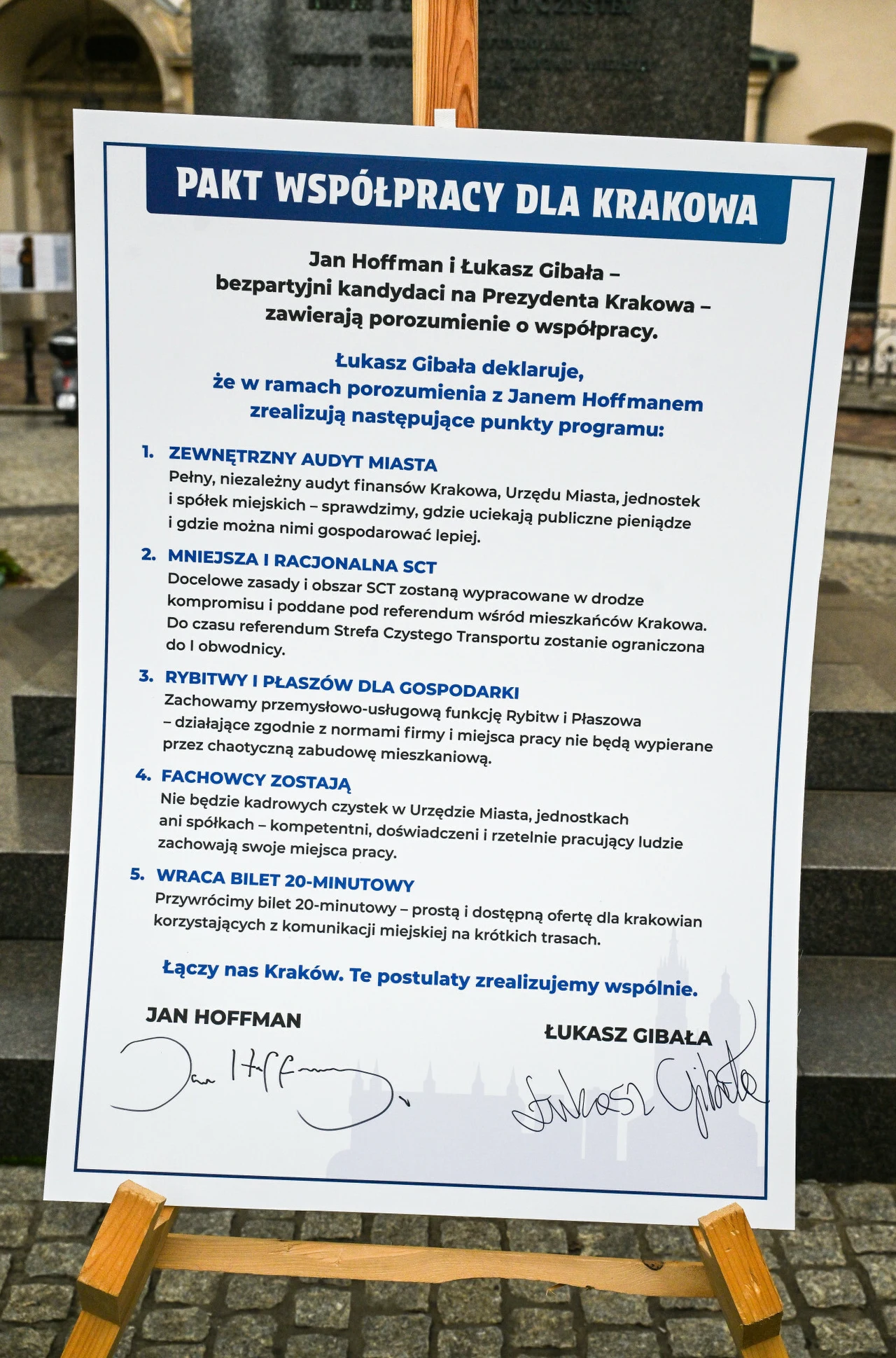 Tablica z podpisanym paktem współpracy dla Krakowa, zawierająca wypunktowane postulaty dotyczące audytu miasta, komunikacji, zieleni i usług, opatrzona podpisami Jana Hoffmana i Łukasza Gibały, ustawiona na sztaludze na zewnątrz.