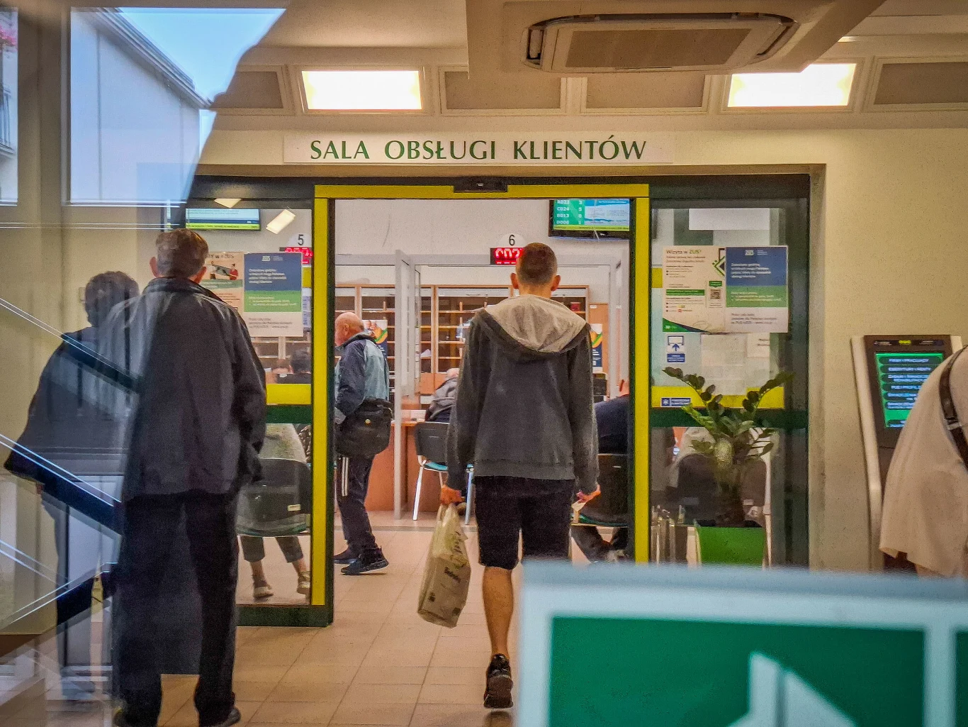 W Polsce rośnie liczba zawieranych umów o dzieło. Osoby wchodzące do sali urzędu, automaty do numerków i tablice informacyjne przy wejściu.