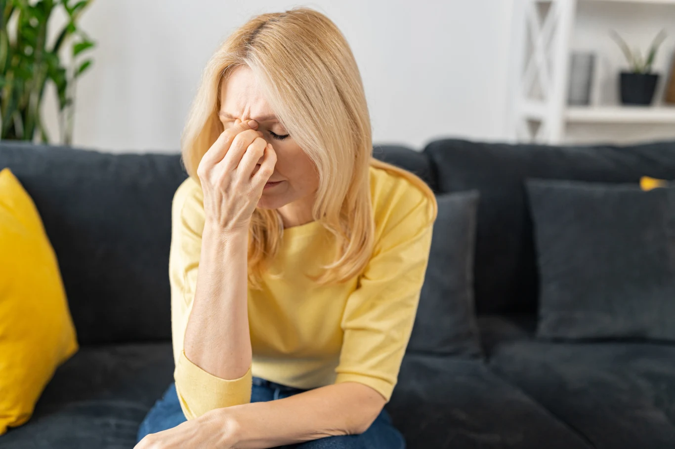 Hormonalna terapia zastępcza nie jest jedyną opcją radzenia sobie z objawami menopauzy. Co mówią badania? Kobieta w jasnożółtej bluzce siedzi na ciemnej sofie, trzyma się za nos i czoło z zamkniętymi oczami.