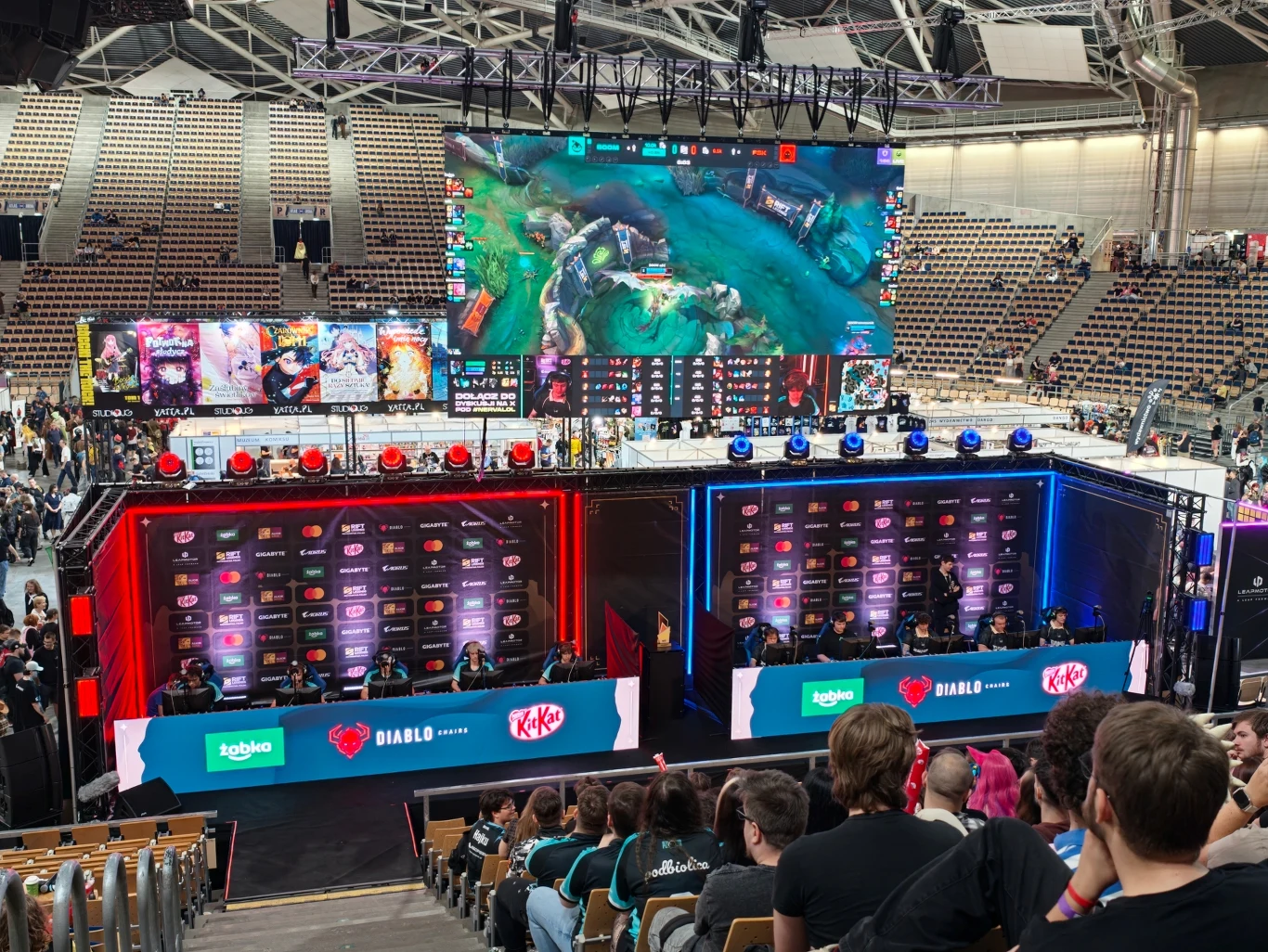 Dwie drużyny e-sportowe rywalizujące ze sobą na dużej arenie, gigantyczny ekran nad sceną wyświetlający rozgrywkę gry komputerowej, w tle widownia śledząca przebieg turnieju.