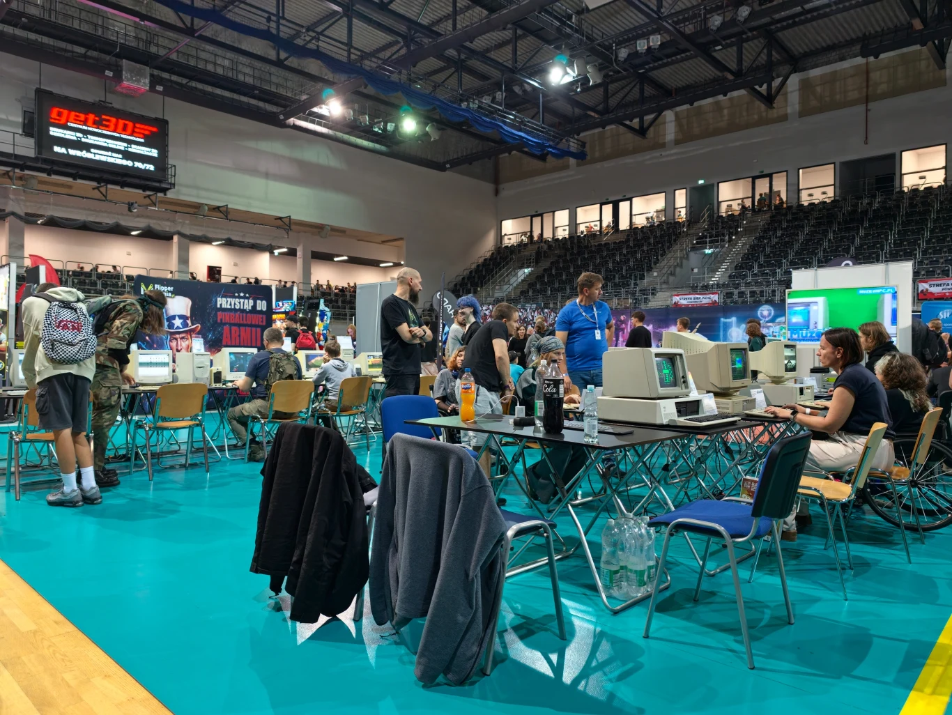 Grupa osób skupiona przy stołach z klasycznymi komputerami, monitorami CRT i klawiaturami, otoczona atmosferą targów lub zlotu komputerowego w dużej hali sportowej.