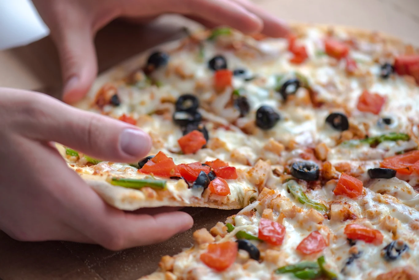 Kawałek pizzy z ciągnącym się serem, oliwkami, papryką i kawałkami kurczaka podnoszony przez dwie ręce.