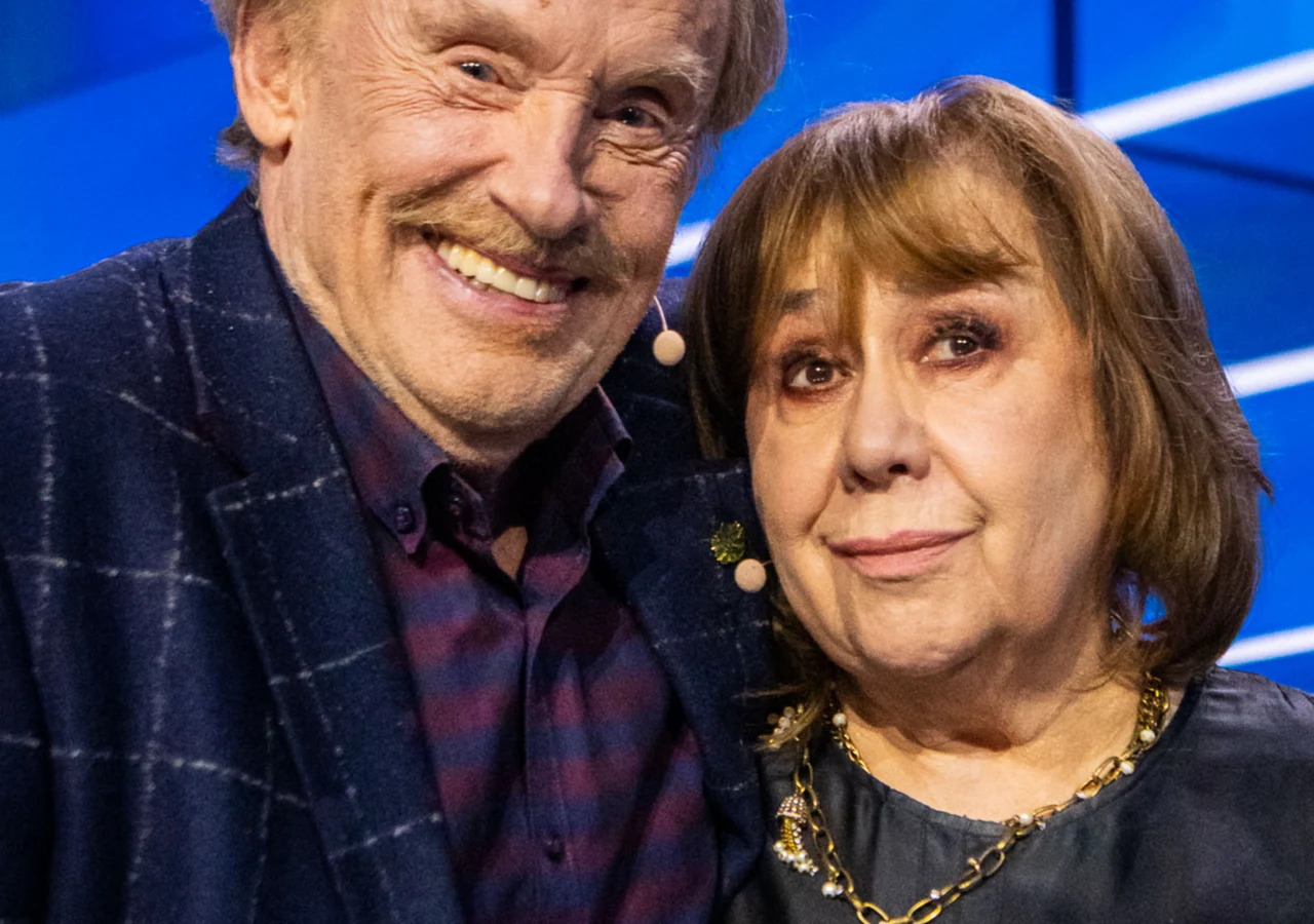 Daniel Olbrychski, Krystyna Demska-Olbrychska Dwie starsze osoby stojące blisko siebie, uśmiechnięty mężczyzna w ciemnym, kraciastym marynarce i kobieta w granatowej bluzce z naszyjnikiem na szyi, oboje mają mikrofony przypięte do twarzy, na jasnoniebieskim tle ze skośnymi liniami.