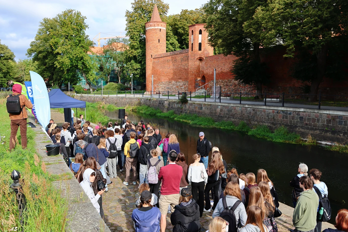 Duża grupa ludzi zgromadzona przy kanałku wodnym naprzeciwko zabytkowych, ceglanych murów z charakterystyczną wieżą, większość osób skierowana twarzą w stronę prowadzącego wydarzenie mężczyzny w ciemnej kurtce.