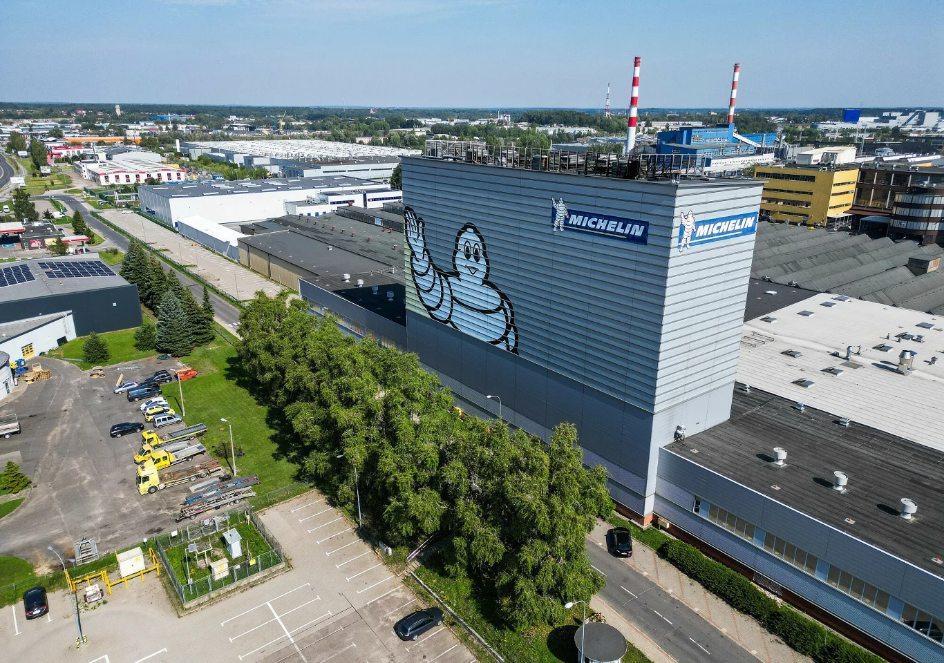 Fabryka Michelin w Olsztynie ruszyła z nową linią produkcyjną Fabryka Michelin w Olsztynie z nową linią produkcyjną, logo Michelin, budynek i kominy w tle.