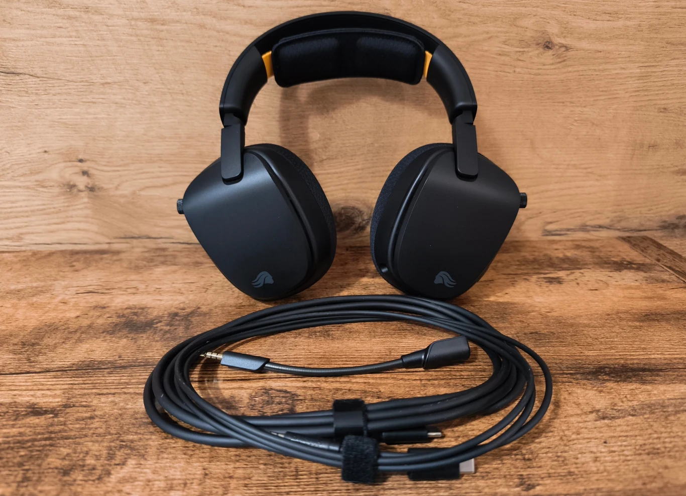 Glorious GHS Eternal RGB Czarne nauszne słuchawki z grubym pałąkiem ustawione pionowo, przed nimi długi przewód audio zakończony wtyczką jack, całość na drewnianej powierzchni.