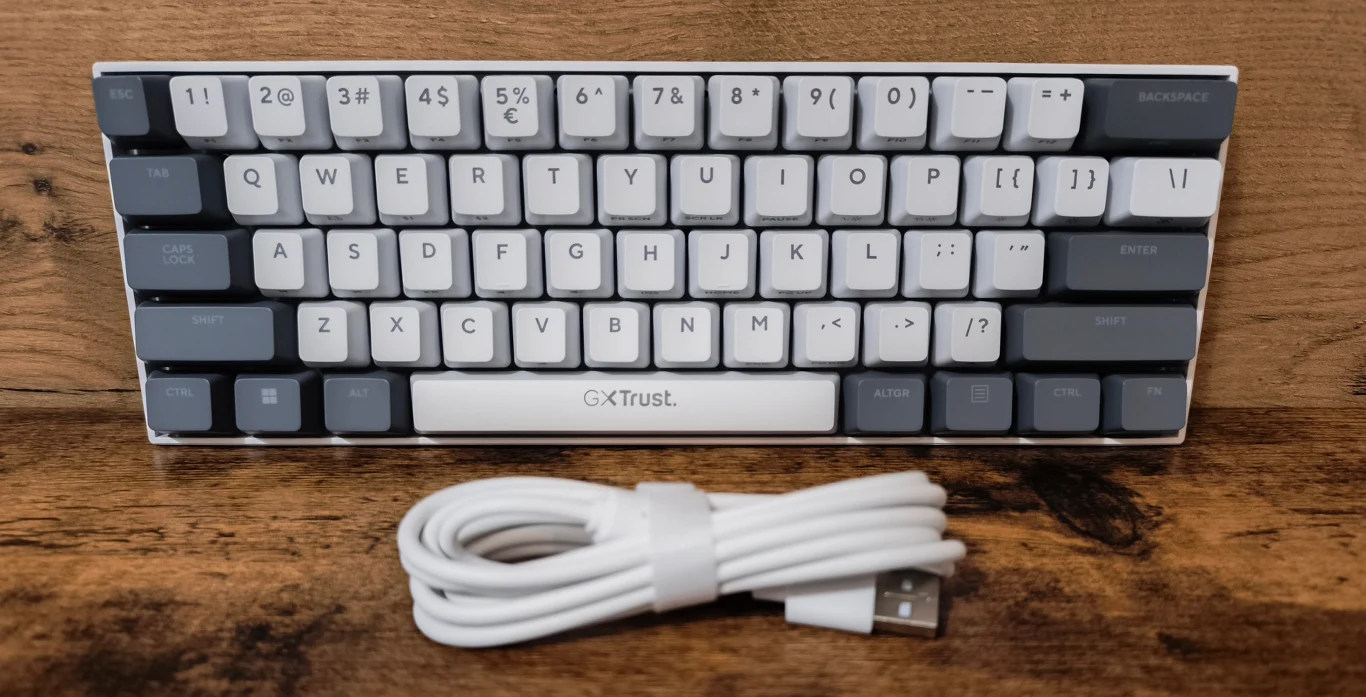 Klawiatura mechaniczna o kompaktowym układzie z ciemnoszarymi i białymi klawiszami oraz zwinięty biały kabel USB umieszczony przed nią na drewnianej powierzchni.