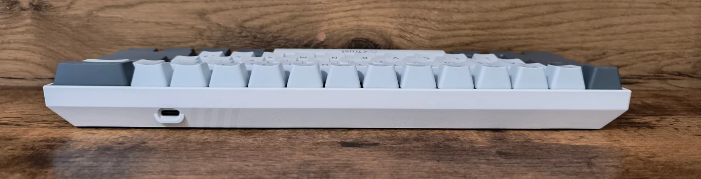 Biała klawiatura mechaniczna z podwyższonymi klawiszami, port USB po lewej stronie obudowy, całość ustawiona na drewnianej powierzchni.