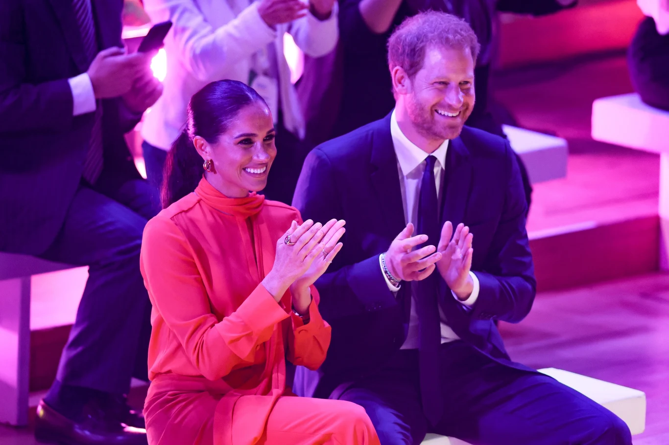 Media wciąż czekają na pierwsze oficjalne wystąpienie Meghan w Wielkiej Brytanii Kobieta w czerwonym stroju i mężczyzna w garniturze siedzą obok siebie na jasnych ławkach, oboje uśmiechnięci i klaszczący, otoczeni przez innych uczestników wydarzenia.