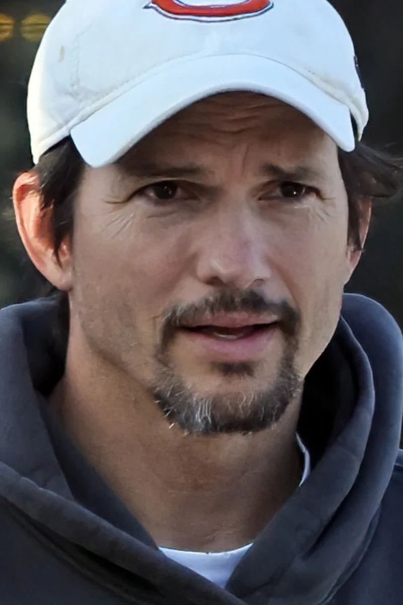 Ashton Kutcher w nowym wydaniu. Aktor zapuścił niewielką bródkę i wąsy Mężczyzna o ciemnych włosach i lekko siwiejącym zaroście, ubrany w szarą bluzę z kapturem oraz jasną czapkę z daszkiem z literą C.