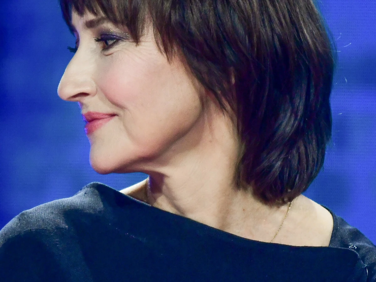 Katarzyna Groniec Portret kobiety o krótkich, ciemnobrązowych włosach i delikatnym uśmiechu, ubranej w ciemną bluzkę na tle niebieskiego, rozmytego tła.